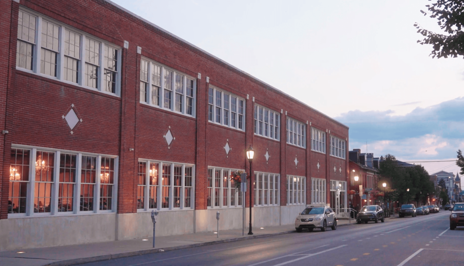 A long, two-story brick building with many rectangular windows lines a city street with parked cars.