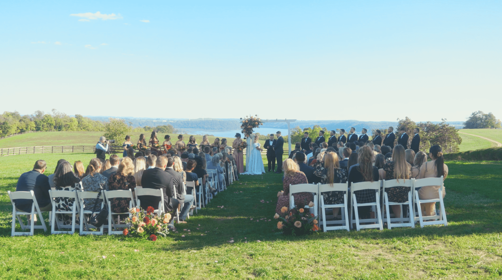 A couple standing at an outdoor wedding ceremony with guests seated in rows on a lawn overlooking a lake.