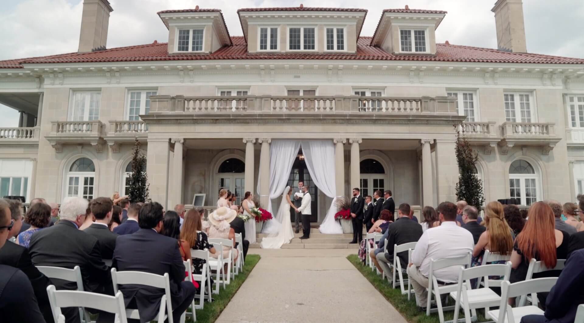 A bride and groom exchange wedding vows in front of a large mansion while guests sit in rows of white chairs.