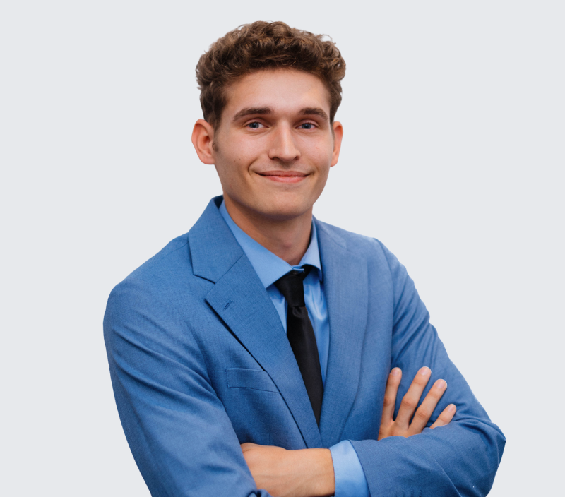 A young man with wavy brown hair wearing a blue suit, blue dress shirt, and black tie, posing with his arms crossed against a light background.