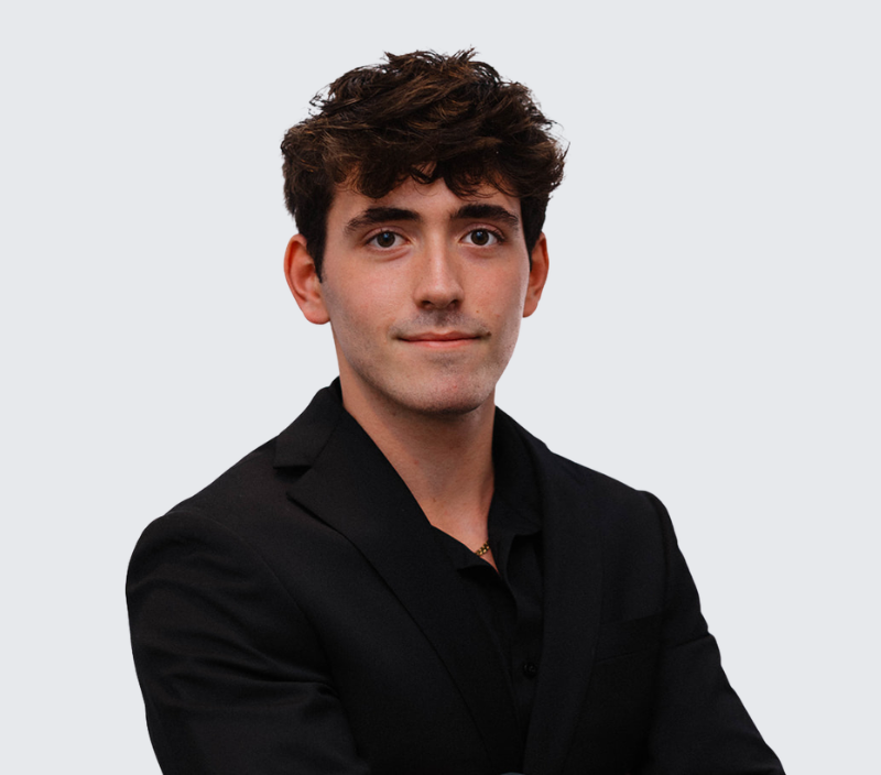 Zain-26 (1) A young man with dark, wavy hair wearing a black blazer and black shirt, centered against a light grey background.