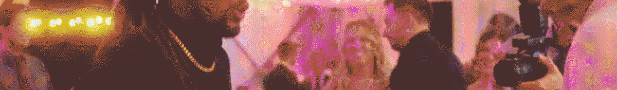 klock-hero-text-bkgd-64 A person with a gold chain speaking to a woman at an event, with a videographer recording them.