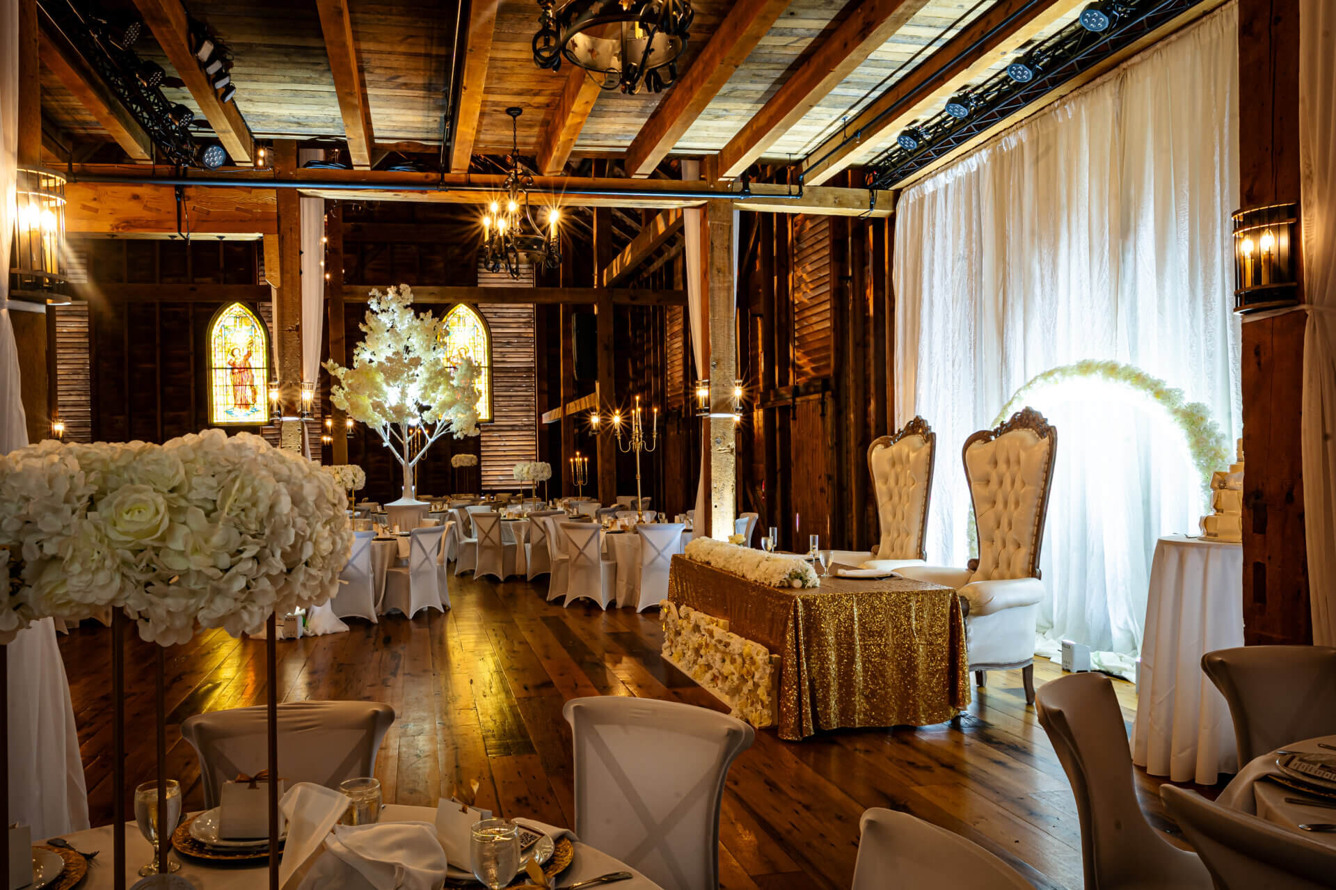 Interior of Stone Gables wedding venue with wooden beams, round guest tables with white floral centerpieces, and a sweetheart table featuring two high-backed chairs and a gold sequin tablecloth.